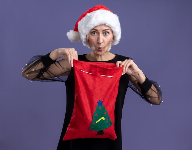 Curious middle-aged blonde woman wearing christmas hat holding christmas sack opening it  with pursed lips isolated on purple wall