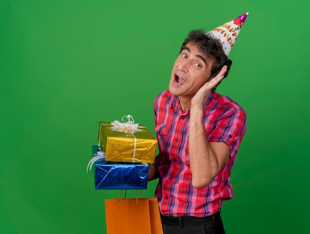 Curioso uomo di mezza età che indossa il cappello di compleanno che tiene confezioni regalo e sacchetto di carta guardando davanti facendo non riesco a sentirti gesto isolato sul muro verde