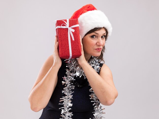 Любопытная женщина средних лет в новогодней шапке и мишурной гирлянде на шее, держащая рождественский подарочный пакет возле головы, смотрит в камеру, изолированную на белом фоне с копией пространства