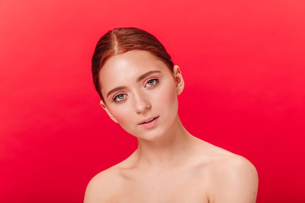 Curious naked girl looking at camera. Studio shot of dreamy ginger woman isolated on red background.