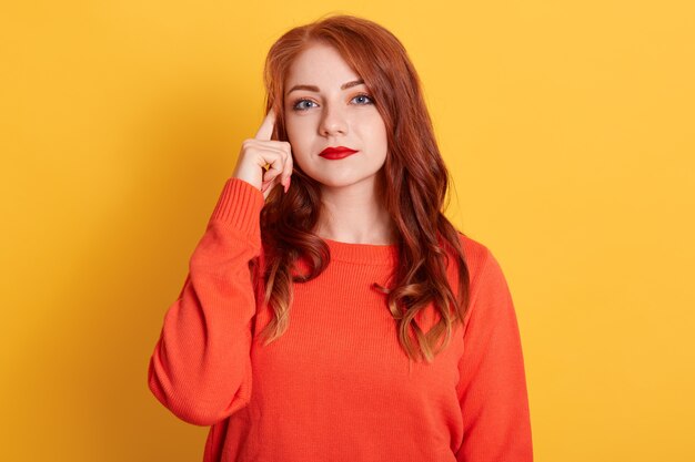 Curious red haired European woman concentrated above, tries to decide something, stands in thoughtful pose, keeps finger near lips, wears orange sweater