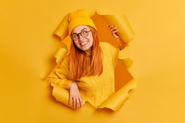 Curious redhead young European woman looks with interest aside has cheerful face expression wears transparent glasses yellow clothes breaks through paper