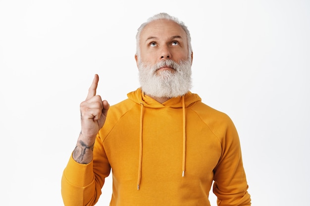 Free photo curious senior man with tattoos look and point up, read promotional text above head, standing in hoodie against white wall