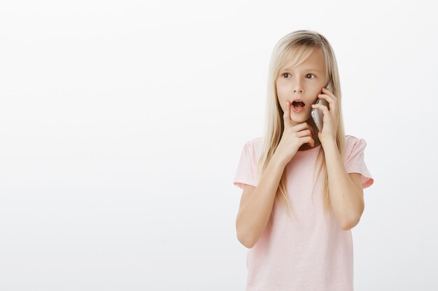 curious smart blond girl in pink t-shirt, talking on smarpthone and holding finger on lip, dropping jaw while listening with interest to speaker