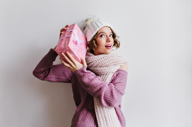 Curious white woman in long knitted scarf holding christmas gift in pink box. Carefree girl in winter hat posing on light background with new year present.