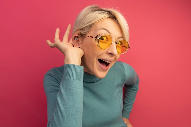 Curious young blonde girl wearing sunglasses keeping hand on waist doing i can't hear you gesture 