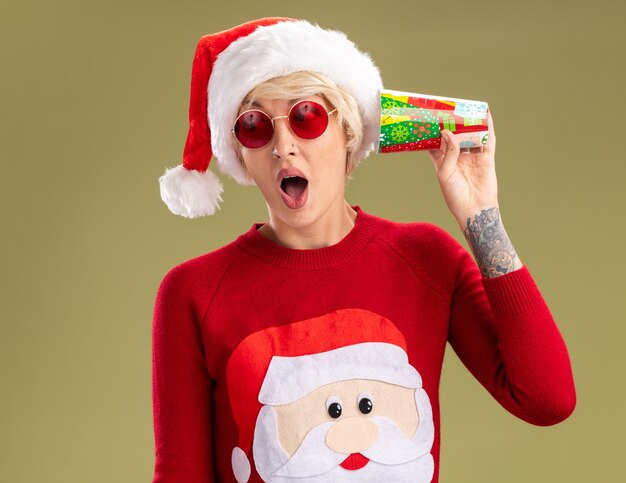curious young blonde woman wearing christmas hat and santa claus christmas sweater with glasses holding plastic christmas cup next to ear listening to secrets looking at side isolated on olive green wall