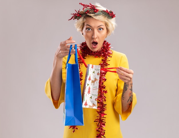 Curious young blonde woman wearing christmas head wreath and tinsel garland around neck holding christmas gift bags opening one looking  isolated on white wall with copy space