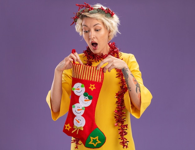 Curious young blonde woman wearing christmas head wreath and tinsel garland around neck holding christmas stocking looking inside it isolated on purple background