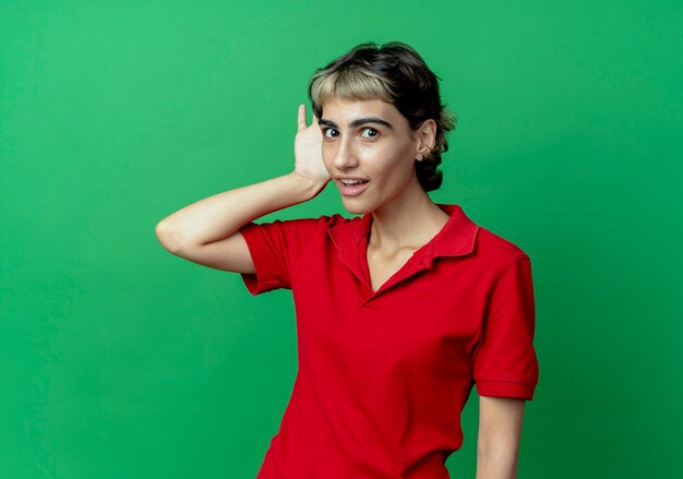 Любопытная молодая кавказская девушка со стрижкой пикси не слышит вашего жеста, изолированного на зеленом фоне с копией пространства