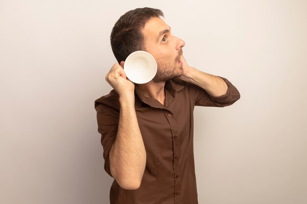 Curious young caucasian man putting hand on face touching ear with cup looking up isolated on white background with copy space