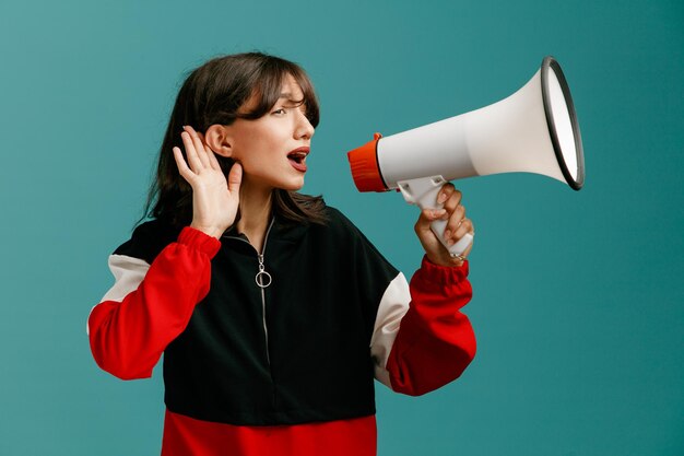 Curious young caucasian woman turning head to side looking at side talking into speaker keeping hand near ear listening isolated on blue background