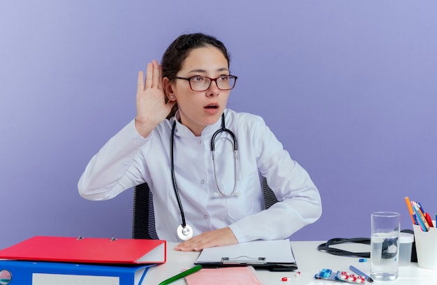 医療用ローブと聴診器を身に着けている好奇心旺盛な若い女性医師が机に座って、医療器具を横から見ていると、ジェスチャーが孤立しているのが聞こえません