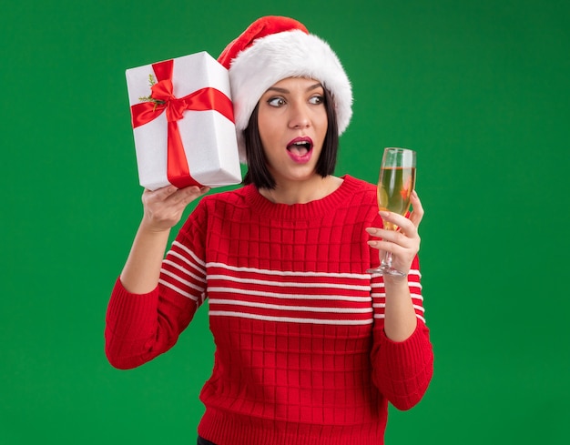 Curious young girl wearing santa hat holding gift package near head and glass of champagne looking at side isolated on green background