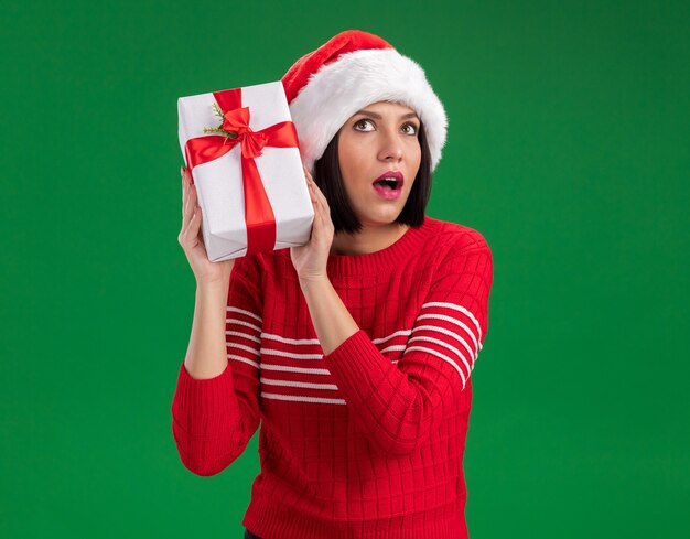 Curious young girl wearing santa hat holding gift package near head looking up listening isolated on green wall with copy space