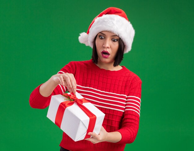 산타 모자를 쓰고 녹색 벽에 고립 된 리본을 잡는 선물 패키지를보고 호기심 어린 소녀