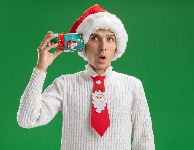 無料Photo クリスマスの帽子とサンタクロースのネクタイを身に着けている好奇心旺盛な若いハンサムな男は、緑の壁で隔離の会話を聞いて側を見て耳の横にプラスチックのクリスマスカップを保持しています