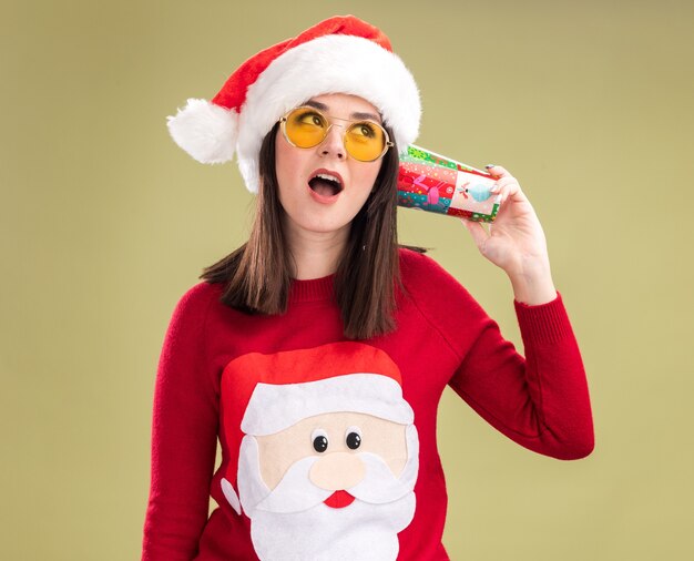 Любопытная молодая симпатичная кавказская девушка в свитере санта-клауса и шляпе с очками держит пластиковый рождественский стаканчик рядом с ушами, слушая разговор