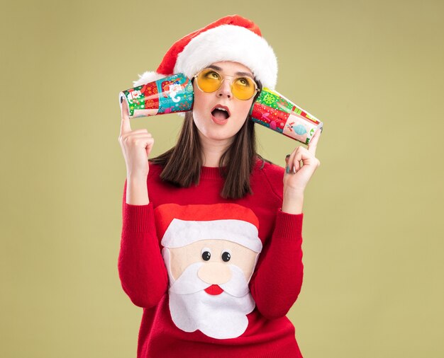 Curious young pretty caucasian girl wearing santa claus sweater and hat with glasses holding plastic christmas cups next to ears listening to conversations 