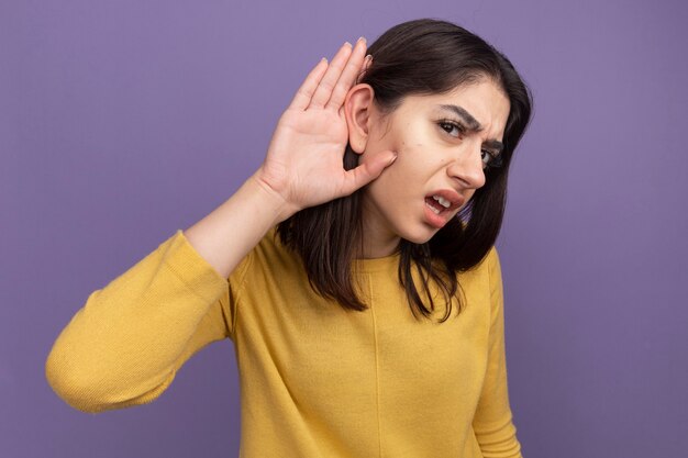 Curious young pretty caucasian woman keeping hand behind ear doing i can't hear you gesture