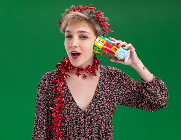 Curious young pretty girl wearing christmas head wreath and tinsel garland around neck holding plastic christmas cup next to ear listening to secrets looking at camera isolated on green background