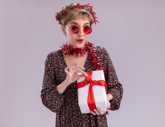 Curious young pretty girl wearing christmas head wreath and tinsel garland around neck with glasses holding gift package grabbing ribbon looking at camera isolated on white background