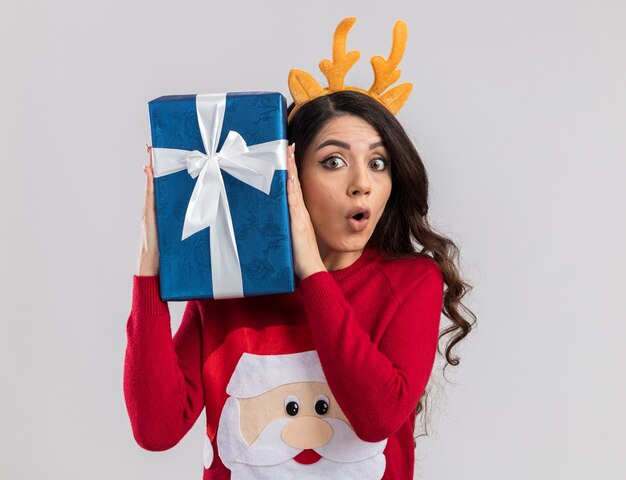 Curious young pretty girl wearing reindeer antlers headband and santa claus sweater holding christmas gift package near head  isolated on white wall with copy space