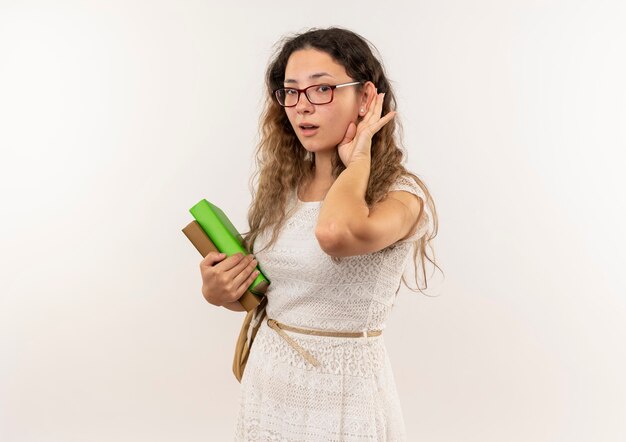 Curious young pretty schoolgirl wearing glasses and back bag holding books doing can't hear you gesture isolated on  with copy space