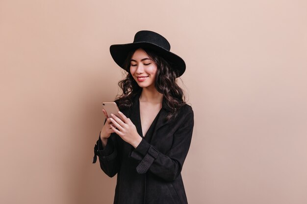 Curly asian woman holding smartphone. Laughing korean woman in coat posing with gadget.