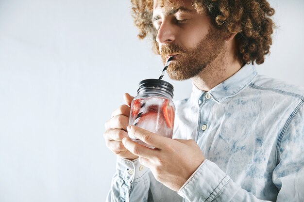 Curly bearded man in shirt enjoys fresh home made strawberry with ice sparkling lemonade through striped drinking straw from rustic transparent jar in hands