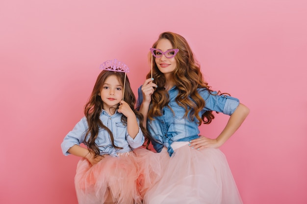 Curly beautiful girl with purple crown posing next to young attractive mother holding masquerade mask on pink background. Adorable woman in vintage outfit having fun with daughter at birthday party.