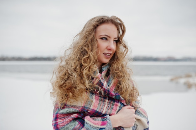 Curly blonde girl in checkered plaid against frozen lake at winter day