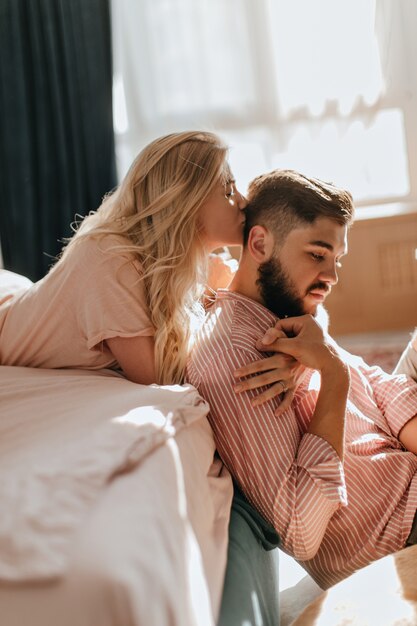 Curly blonde girl kisses her boyfriend in striped shirt sitting on floor in bright bedroom.