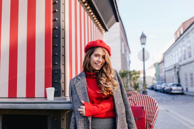 Curly brown-haired girl with sincere smile posing in gray coat on beautiful european street
