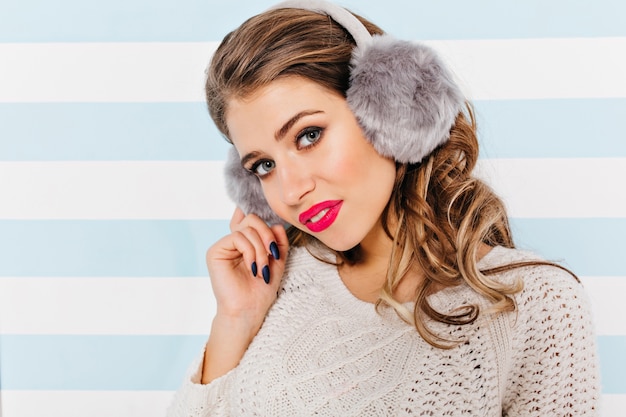 Curly brown-haired woman in white knitted sweater looks cute and shy, posing for close-up portrait