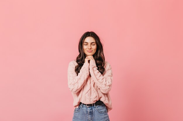 Curly brunette calmly smiles with closed eyes on pink background and makes wish.