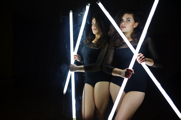 Curly brunette plus size model with two long led lamp at dance room with mirror