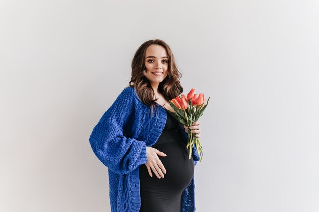 Curly brunette pregnant woman in black dress and blue cardigan holds tulips. Pretty lady smiles on white background.