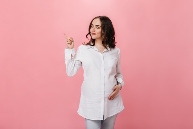 Curly brunette pregnant woman in white shirt pointing to place for text on isolated. Happy girl in denim pants smiles and poses on pink background.