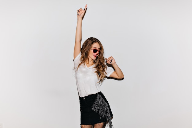 Curly brunette woman in sunglasses looking away while dancing on white wall