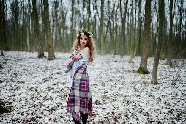 Curly cute blonde girl with wreath in checkered plaid at snowy forest in winter day