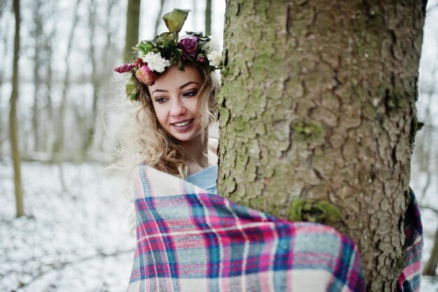 Curly cute blonde girl with wreath in checkered plaid at snowy forest in winter day