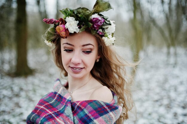 Curly cute blonde girl with wreath in checkered plaid at snowy forest in winter day