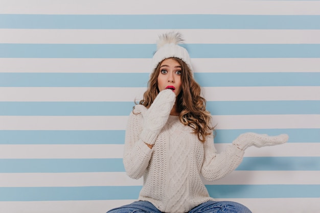 Foto gratuita ragazza bionda scura riccia in sorpresa copre il viso con le mani. ritratto di signora in maglione lavorato a maglia bianco