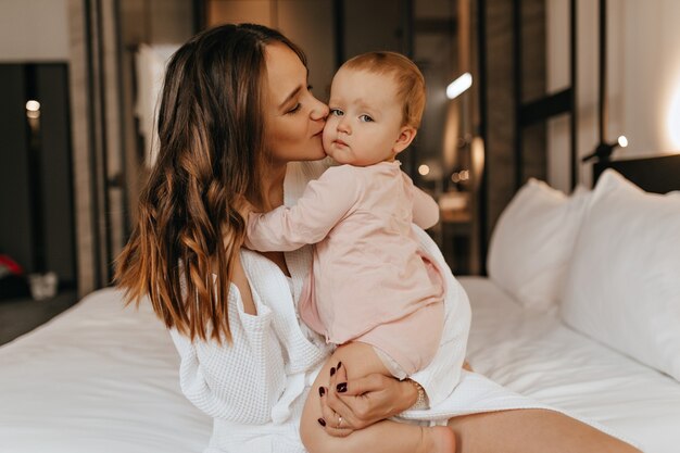 Curly dark-haired woman lovingly kisses her little daughter. Shot of young mother in white coat and her kid in bedroom.