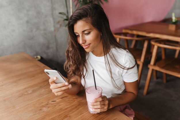 Curly dark-haired woman in white T-shirt writes message in phone and holds milkshake while sitting in cafe