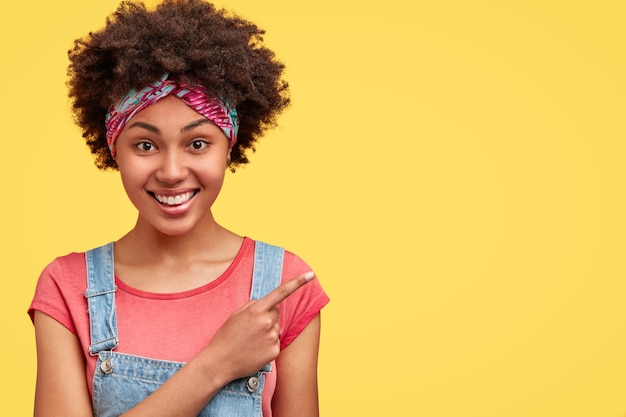 Curly dark skinned female with joyful expression, dressed casually, points at upper right corner, isolated over yellow wall, suggests to visit this cafe. Positive African American woman indoor