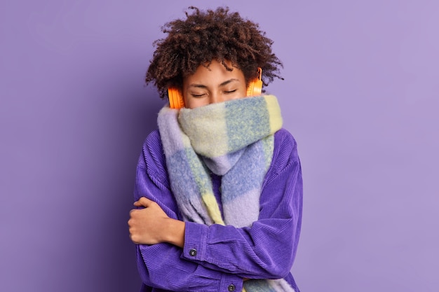 Бесплатное фото Кудрявая этническая девочка-подросток пытается согреться на прогулках в холодную погоду в куртке и теплом шарфе на шее с закрытыми глазами обнимает собственное тело позирует на фоне ярко-фиолетовой стены наслаждается музыкой