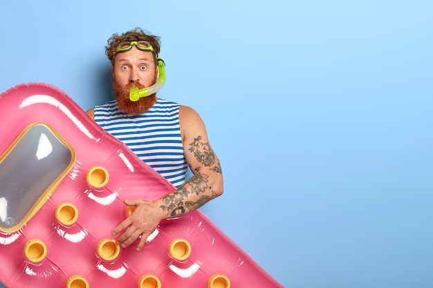 Foto gratuita l'uomo dai capelli rossi ricci indossa occhiali da nuoto, maschera per lo snorkeling e materasso rosa gonfiato, pronto per le immersioni in acqua di mare, indossa un giubbotto blu e bianco a strisce