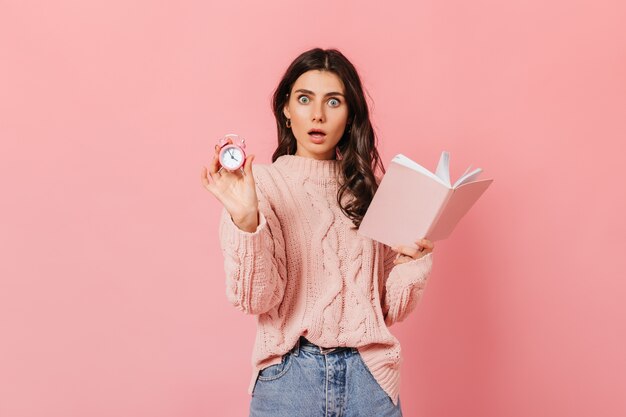 Curly girl in amazement looks into camera on pink background. Woman in sweater holding book and alarm clock.